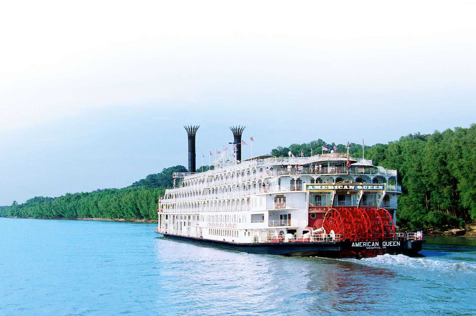 Historisches Mississippi-Flussdampfschiff "American Queen" auf dem Wasser, umgeben von grünen Uferbäumen, symbolisiert amerikanische Flusskreuzfahrten und Tourismus in den USA.