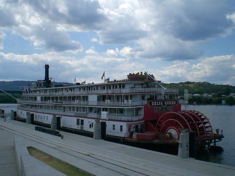 Chattanooga Luxus-Passagierdampfschiff am Ufer in den USA, bekannt als "Delta Queen", mit historischem Rad auf der Rückseite, auf Wasser mit Stadt- und Hügellandschaft im Hintergrund, ideal für USA-Reisebilder.