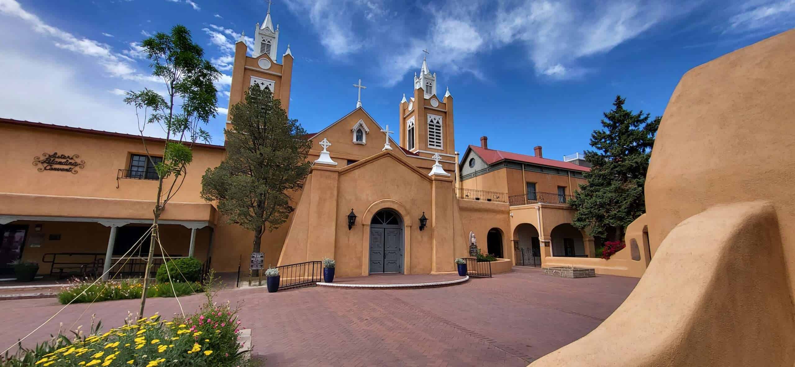 Historische Kirche im südwestlichen Stil mit zwei Türmen und blauen Himmel, umgeben von blühenden Blumen und mediterranen Gebäuden.