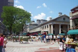 Vielfalt auf dem Bauernmarkt in Boston mit Kunstwerken, Essenständen und Touristen vor historischen Gebäuden und modernen Wolkenkratzern, typisch für die USA.